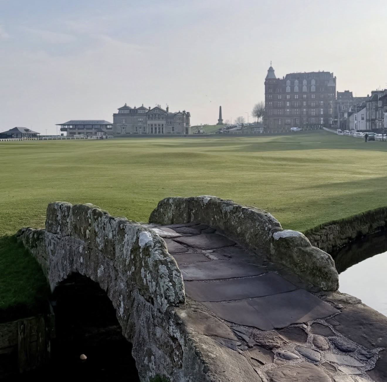 The stone Swilcan Bridge on the Old Course at St Andrews with the R&A clubhouse and Hamilton Grand beyond