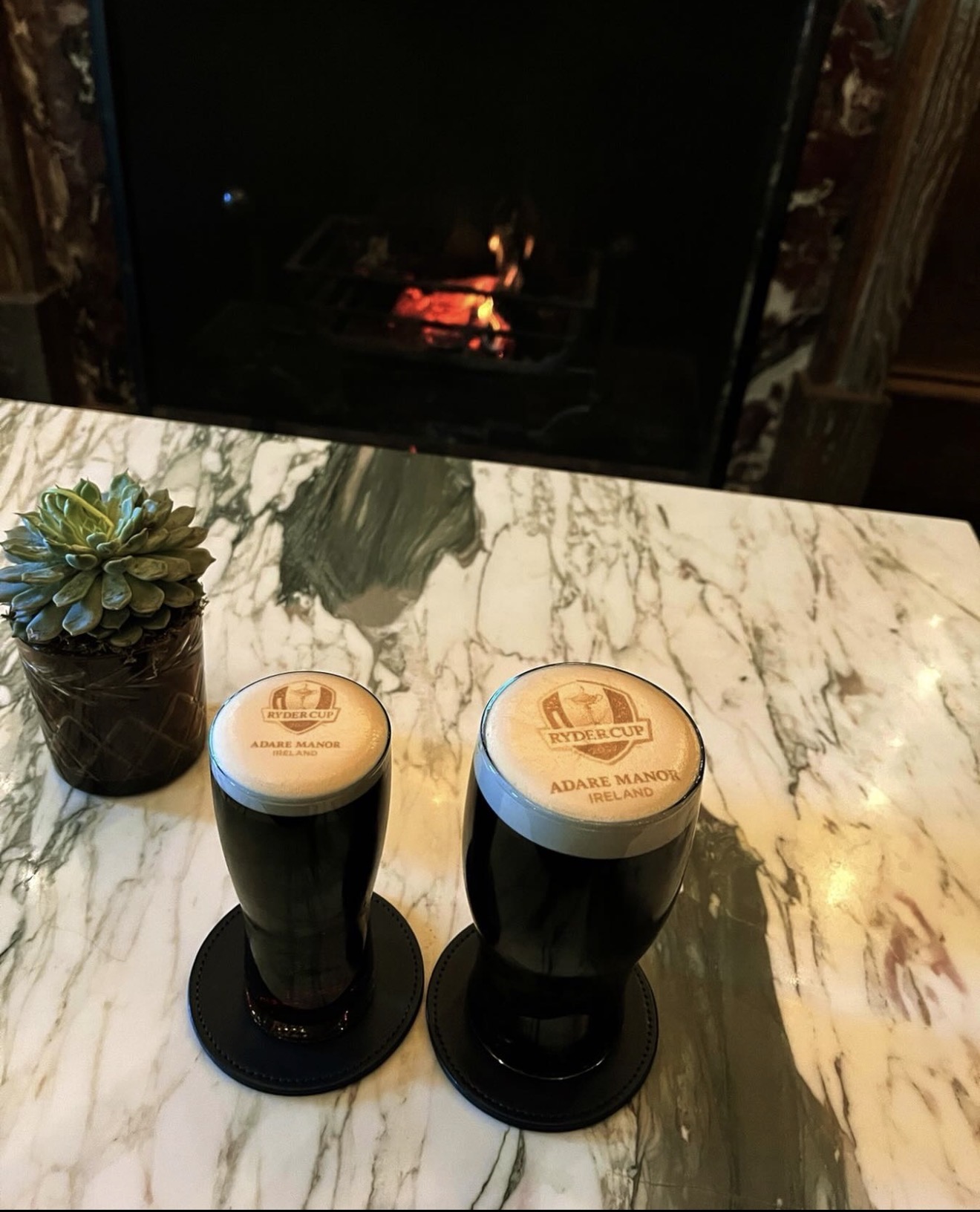 Two Guinness pints with Ryder Cup Adare Manor Ireland logos on a marble table by a fireplace